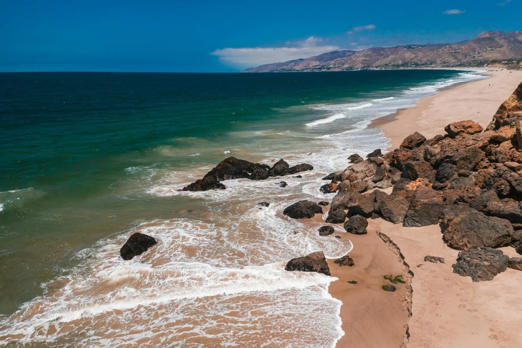 Beautiful Malibu beach coastline with ocean waves, rocky shore, sandy beach, and mountains under a blue sky.